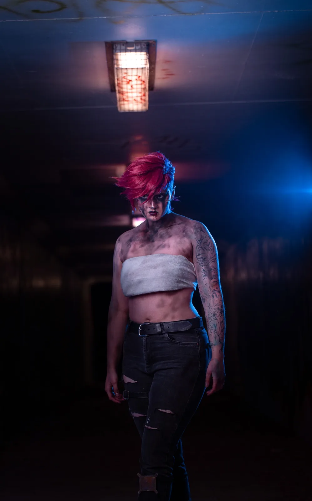 A person with bright red hair and dark makeup stands in a dimly lit hallway, wearing a white crop top and ripped jeans, with…