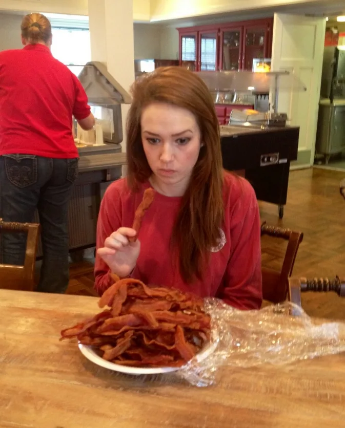 A young woman with reddish-brown hair, wearing a red shirt, sits at a wooden table with a large plate piled high with bacon…