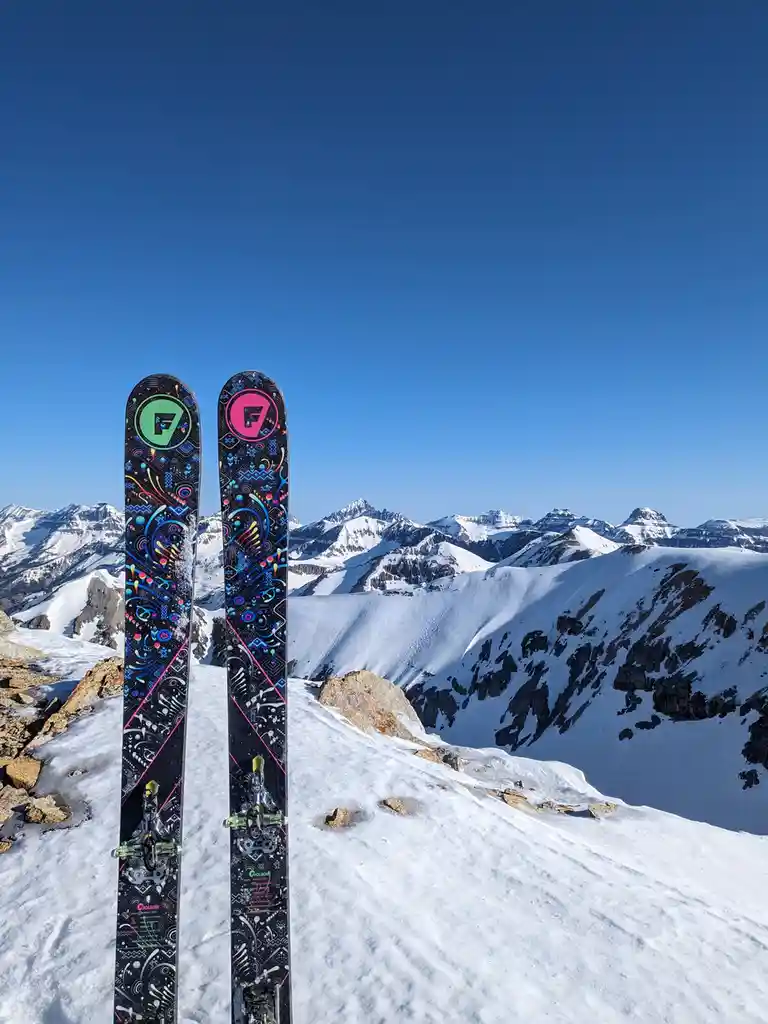 A pair of vibrantly patterned skis with green and pink 'F' logos stand upright in fresh snow on a mountain ridge…