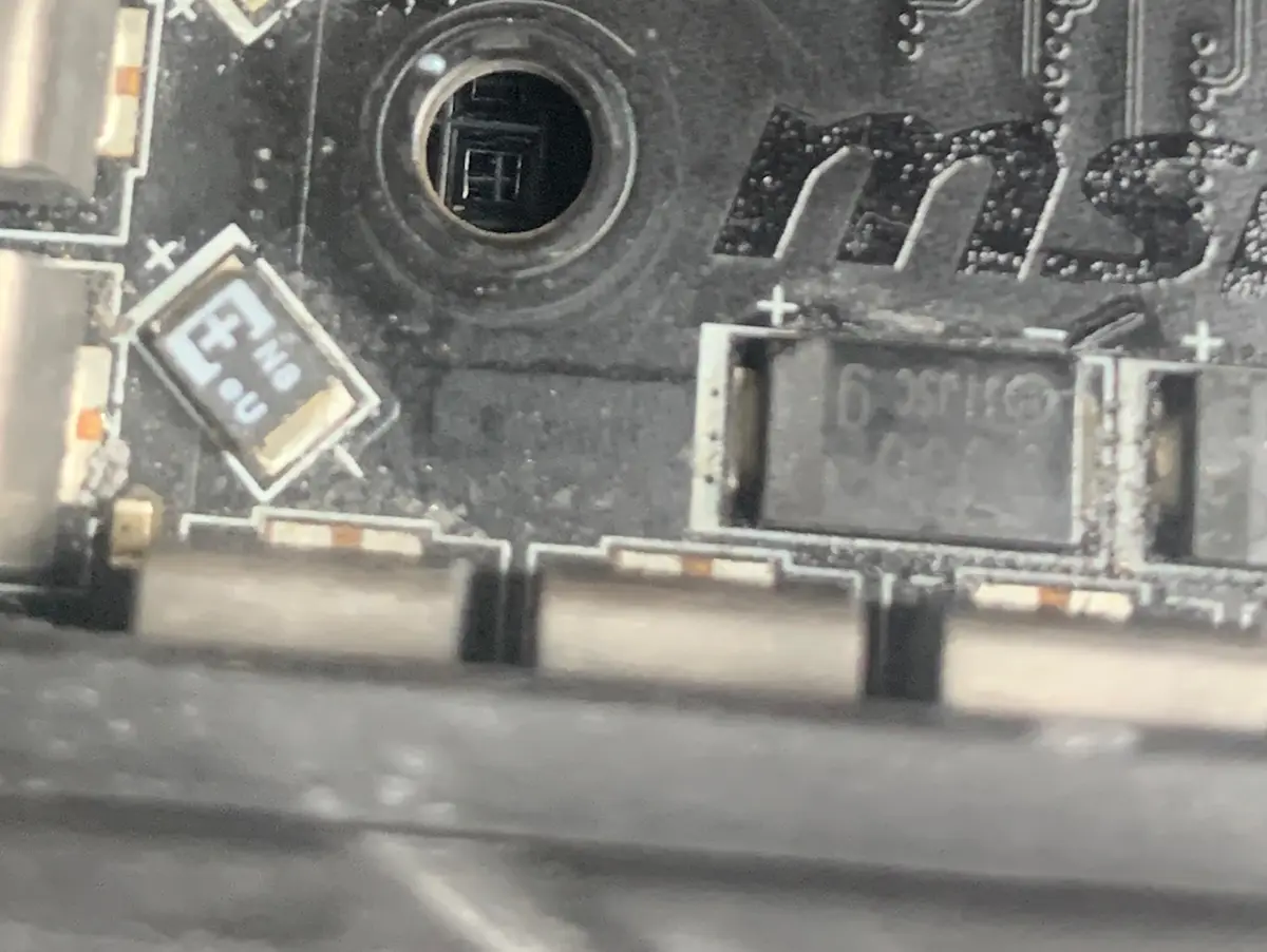 A close-up photo of a damaged computer motherboard with visible burn marks, melted components, and a circular hole near the…