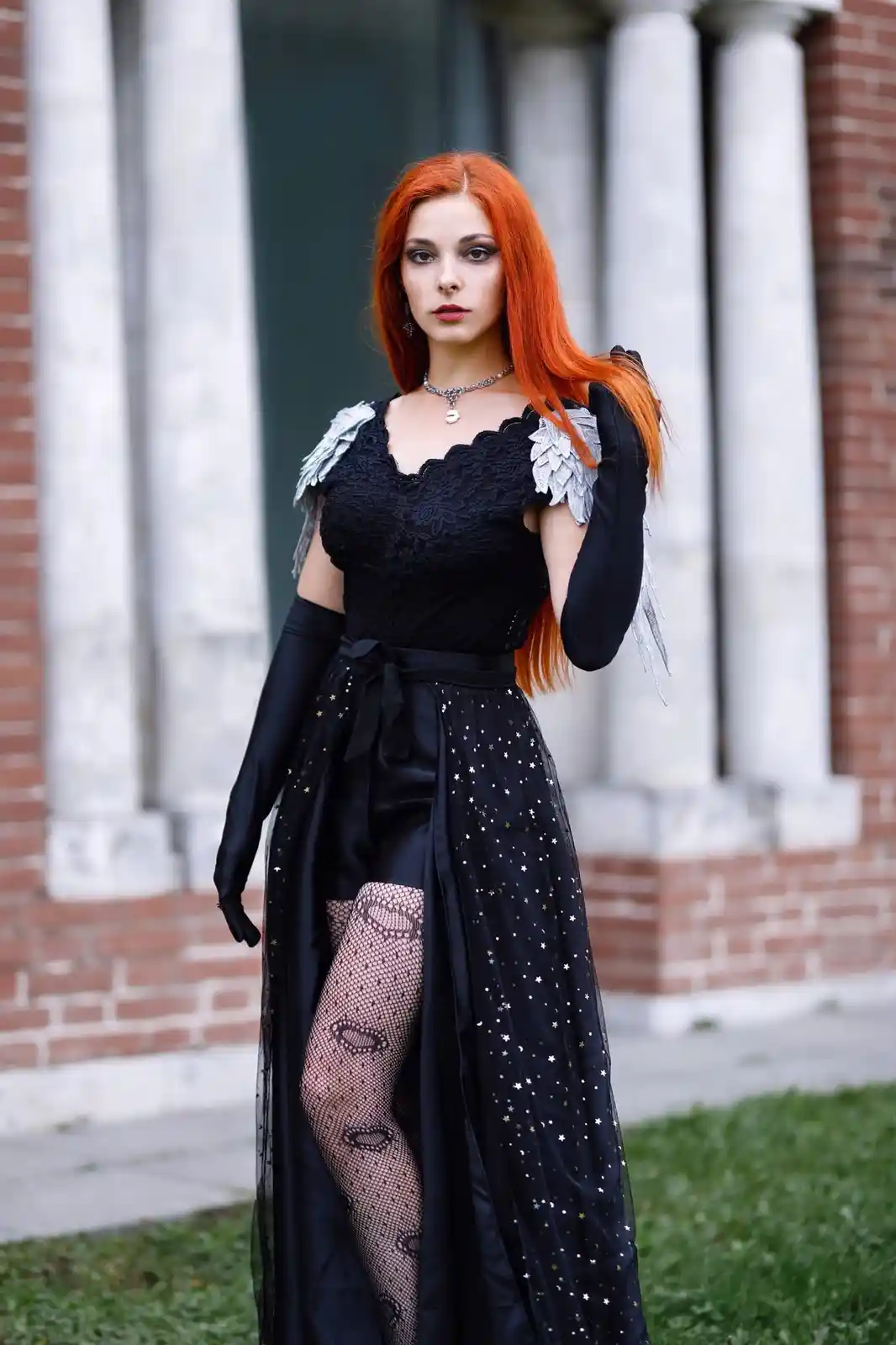 A woman with long orange hair stands outdoors in front of classical columns, wearing a black lace top, a long black skirt…
