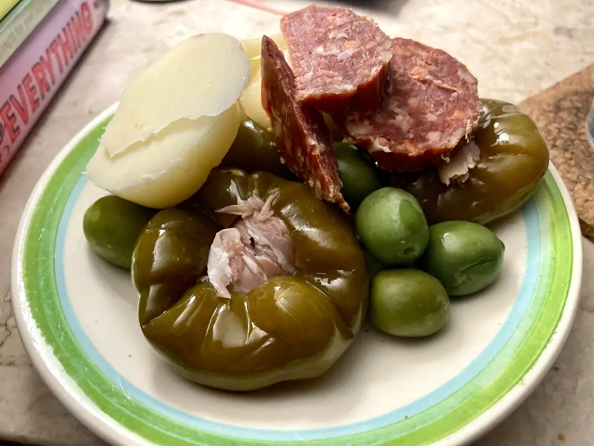 A close-up photo of a plate of tapas featuring sliced salami, green olives, a stuffed green pepper with meat inside, and a…
