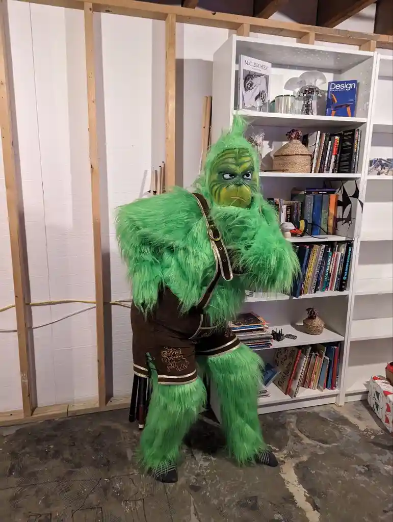 A person in a full-body green Grinch costume, complete with furry texture and brown overalls, stands in a room with…