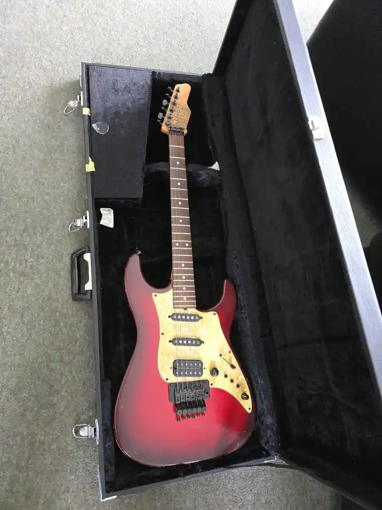 A red and gold electric guitar with a pickguard sits inside an open black hardshell case on a gray carpet. The overlaid text…
