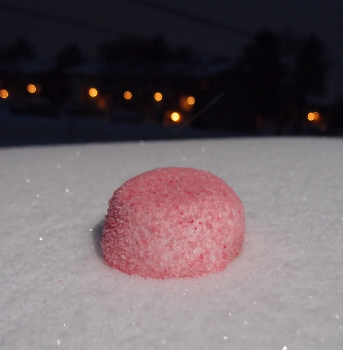 A pink, coconut-covered confection sits in fresh snow with a blurry nighttime background of streetlights and trees. The…