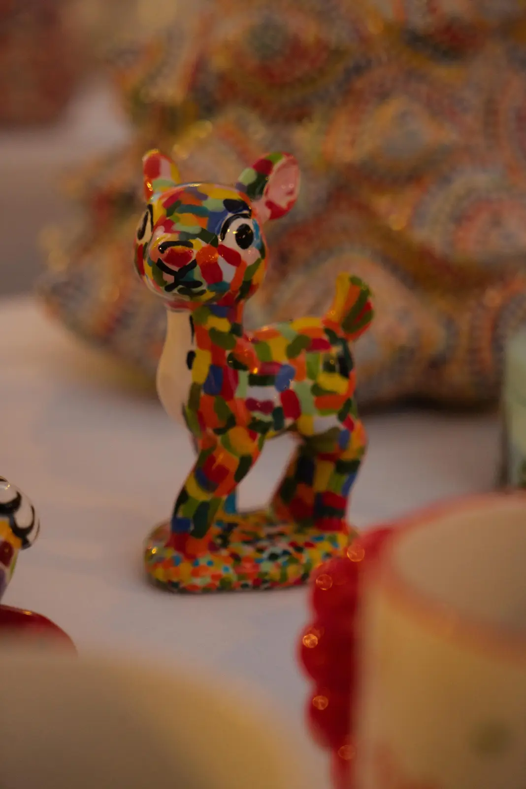A small, colorful ceramic figurine of a deer, covered in multicolored patches, stands on a white surface. The overlaid text…