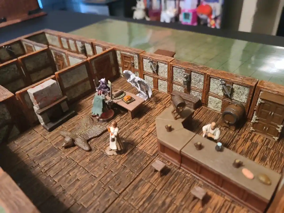 A tabletop miniature scene depicts a fantasy tavern with wooden floors, stone walls, and a bar. Several miniature figures…