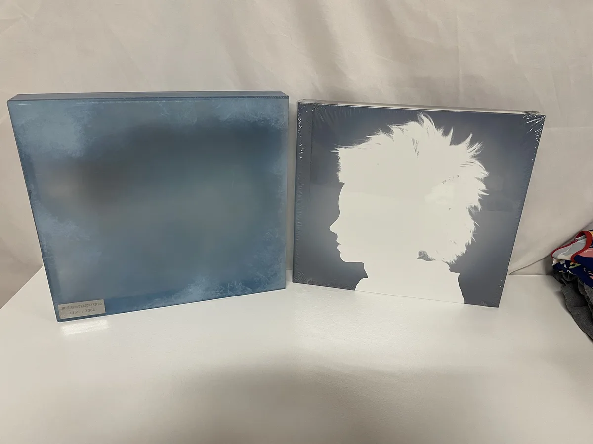 Two sealed, collectible vinyl records for 'The Girl with the Dragon Tattoo' stand side by side on a white surface. The left…