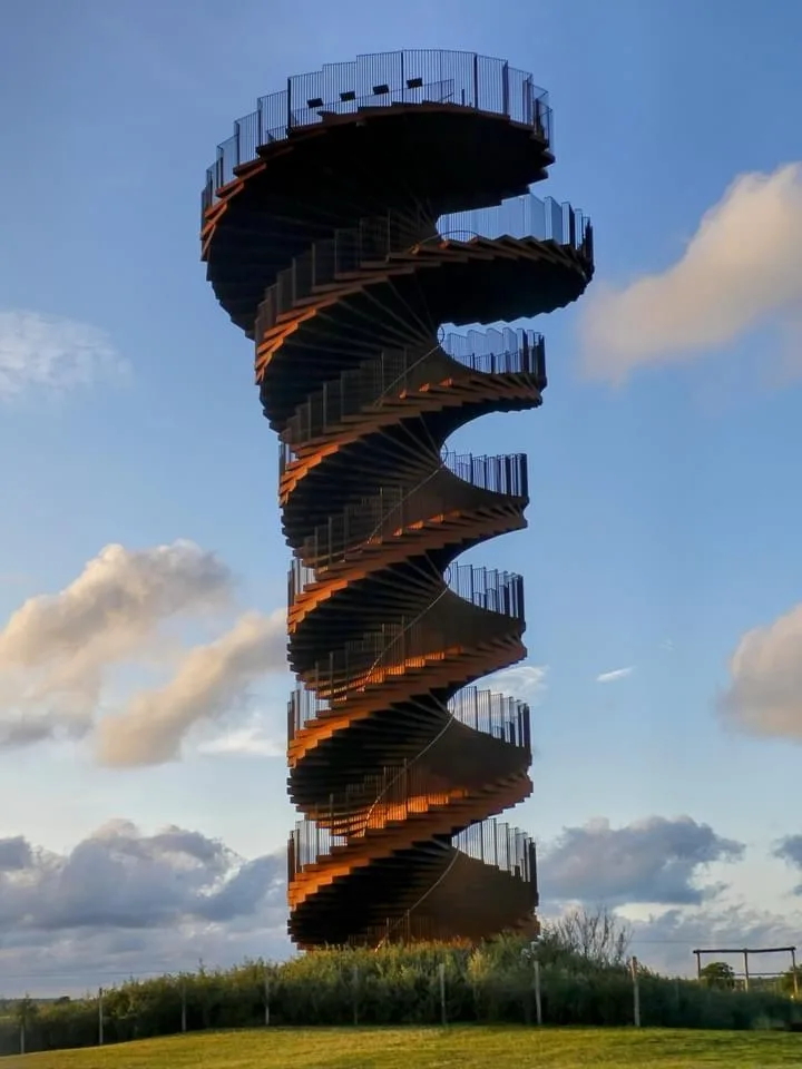 A tall, spiraling metal observation tower with multiple circular platforms and railings, set against a partly cloudy blue…