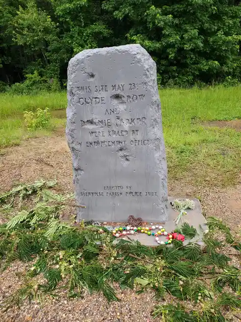 A weathered grey stone memorial in a grassy, wooded area commemorates Clyde Row and Bennie Parker, killed by a law…