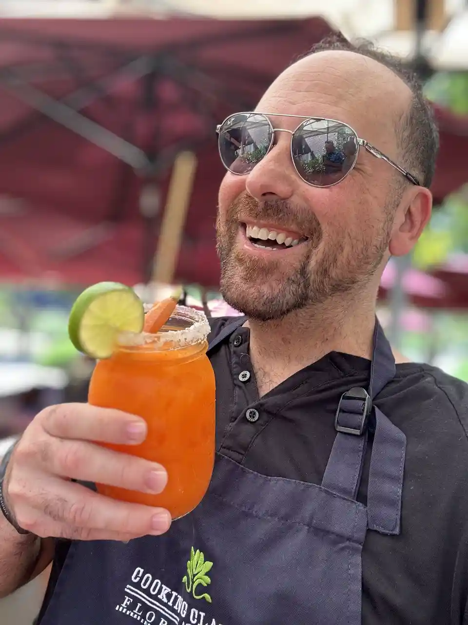 A smiling man wearing sunglasses and a dark apron holds up a bright orange cocktail in a mason jar, garnished with a lime…