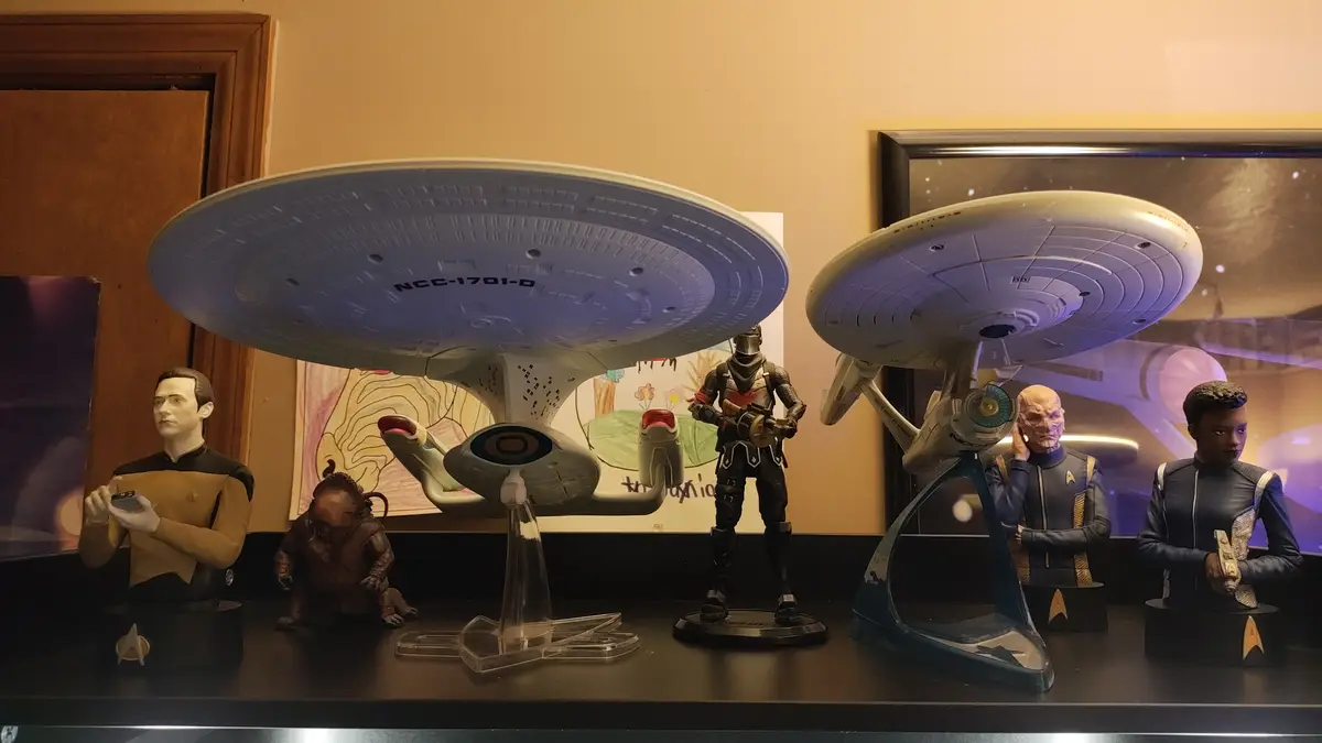 A shelf displays Star Trek collectibles including two starships (one labeled NCC-1701-D) and busts of characters from…