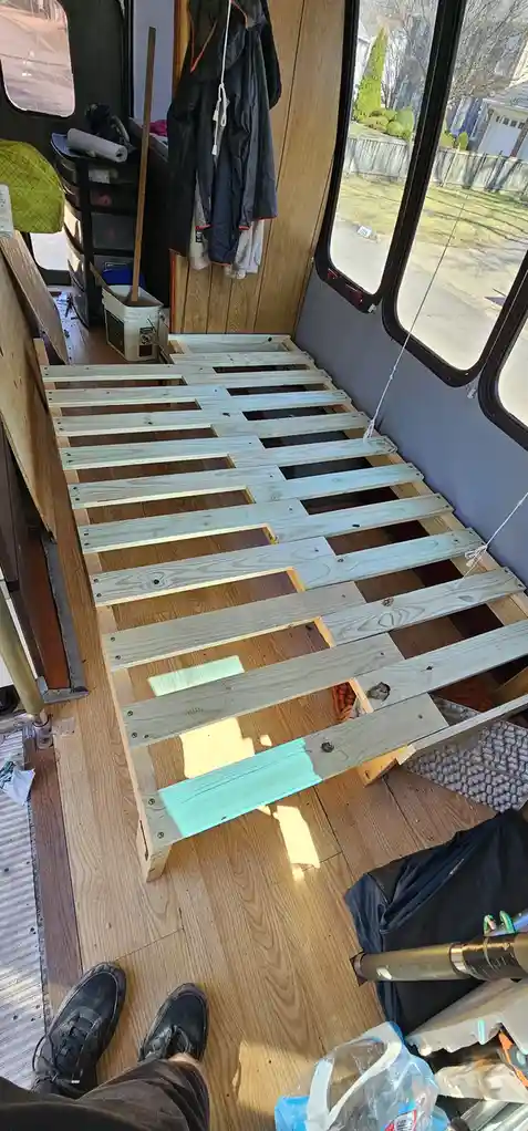 A first-person view inside a van or camper showing a partially built wooden bed frame with slats. One slat near the bottom…