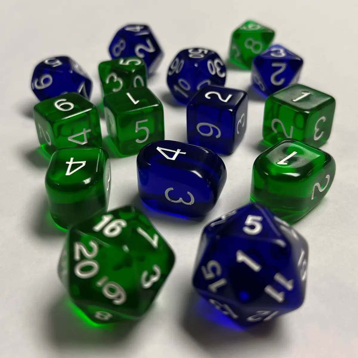 A collection of translucent green and blue polyhedral dice with white numbers are scattered on a white surface, showing…