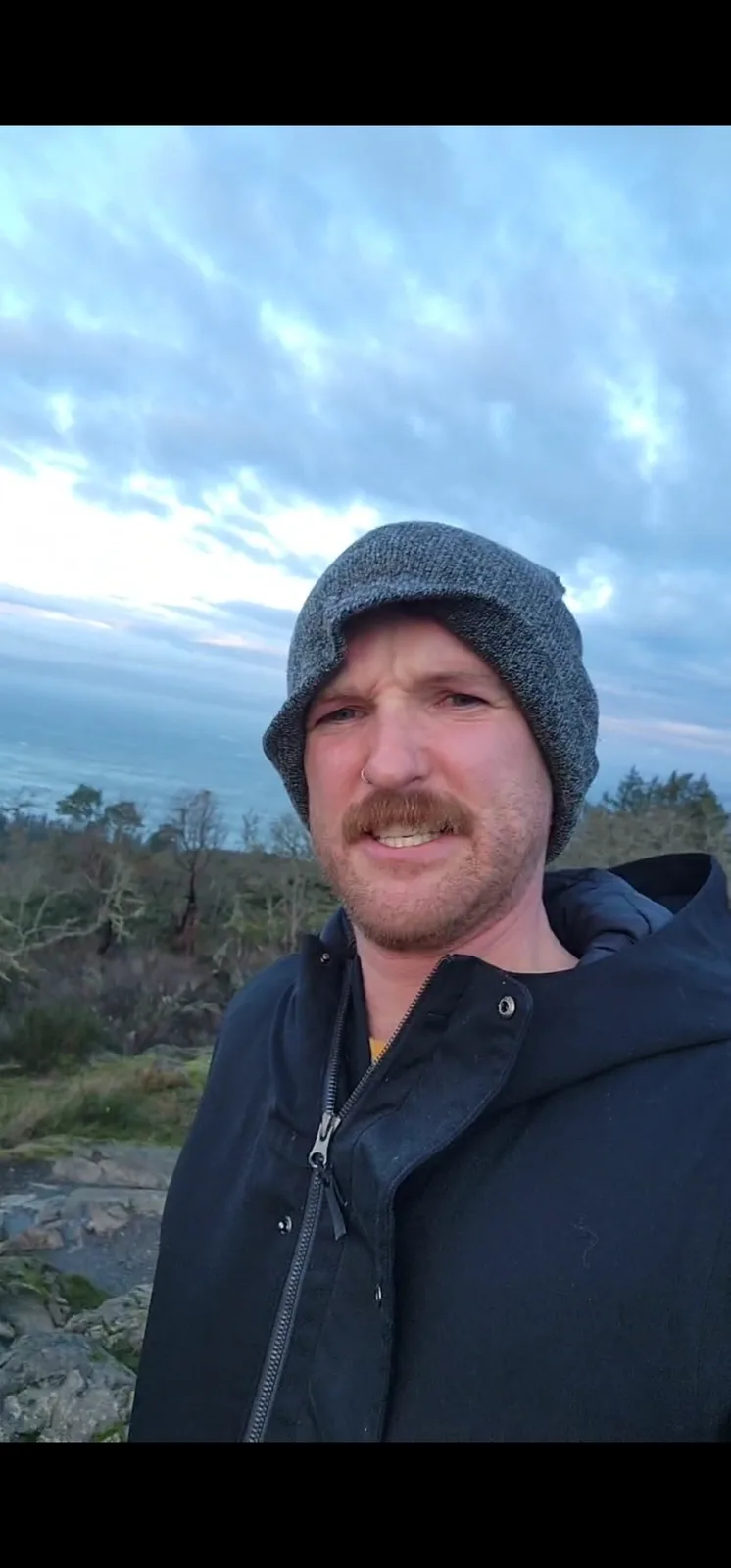 A man with a mustache and a gray beanie stands outdoors in a cold, cloudy environment with trees and rocks behind him. He…