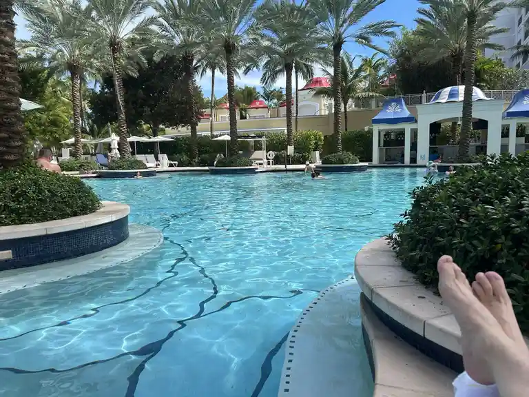 A first-person perspective shows bare feet dangling over the edge of a resort-style swimming pool, with clear blue water…