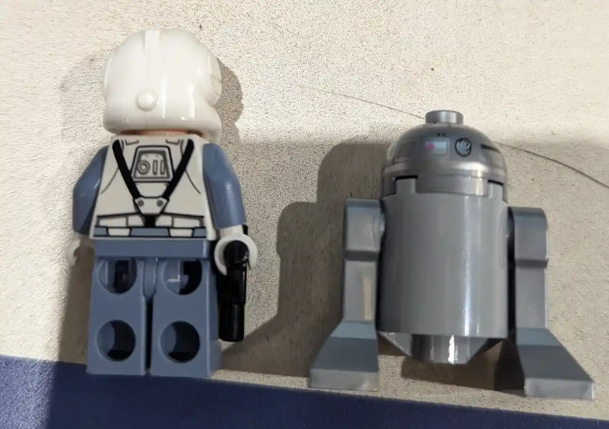 A Lego minifigure of a white-helmeted character, resembling a Star Wars pilot, stands next to a gray Lego R2-D2 droid. Both…