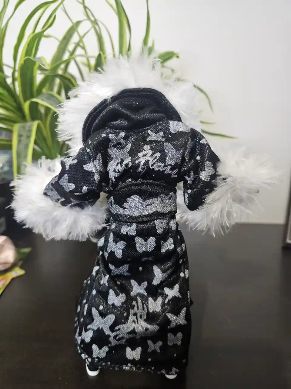A small doll or figurine, viewed from behind, wears a black robe with white butterfly patterns and white faux fur trim on…
