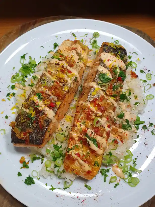 A close-up photo of a white plate holding two grilled salmon fillets resting on a bed of white rice, garnished with chopped…