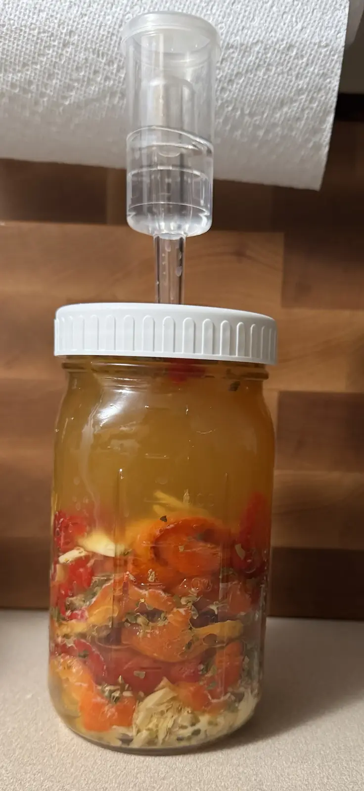 A glass mason jar filled with sliced red and orange peppers, garlic cloves, and herbs submerged in brine, sealed with a…
