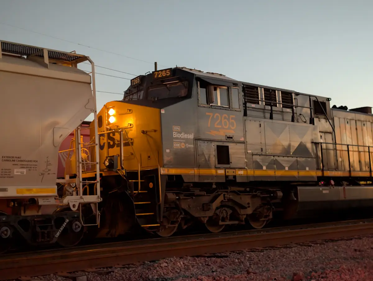 A CSX freight train locomotive, numbered 7265, is shown at dusk with its headlights on, featuring a prominent '20%…