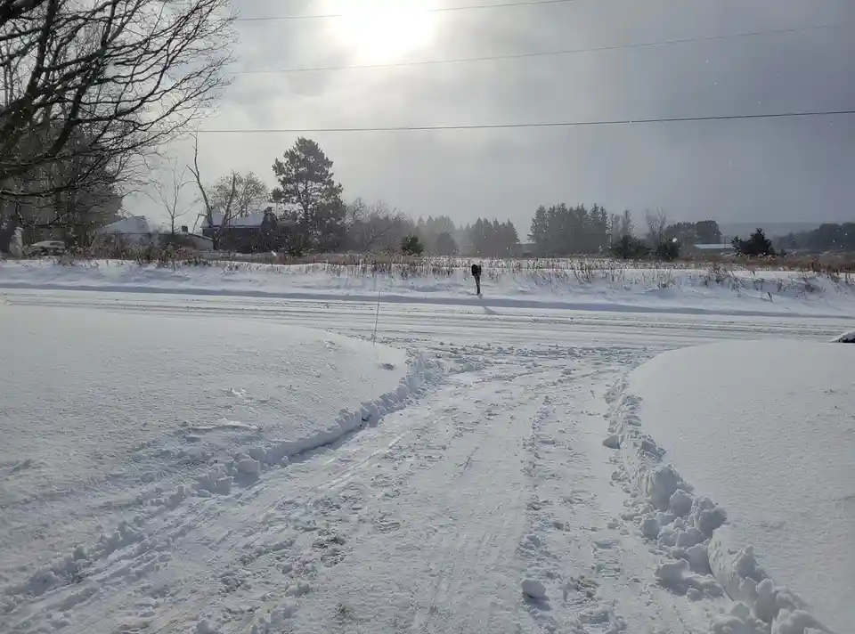 A snowy rural landscape with a person standing in the distance on a snow-covered field, with footprints and tire tracks…