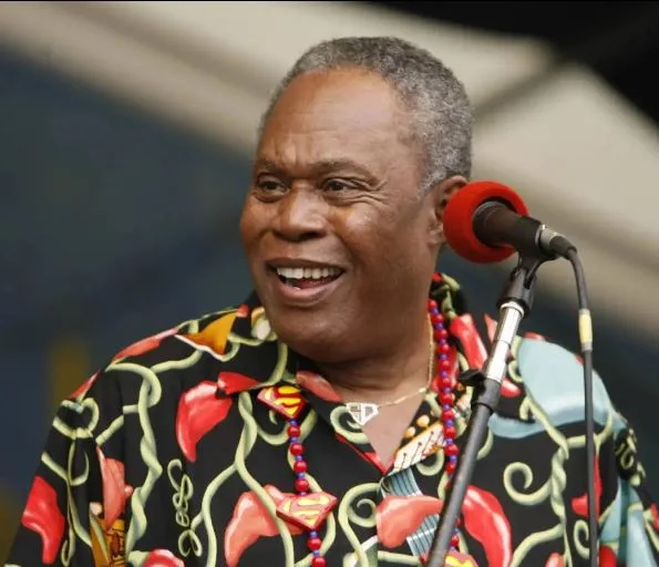 a smiling older black man with gray hair stands at a microphone wearing a black shirt with red and yellow Superman logos and…