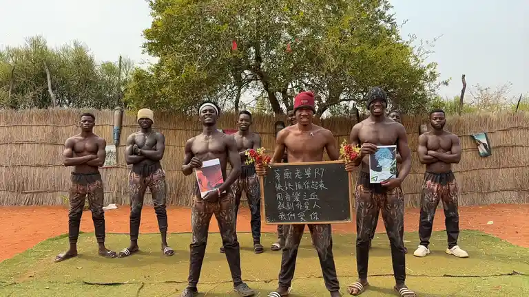 A group of shirtless Black men in matching wolf-print pants stand outdoors on artificial grass in front of a thatched fence…