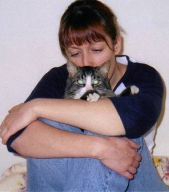 A woman in a dark blue shirt and jeans is hugging a tabby cat tightly, her face pressed against the cat's head. The cat has…