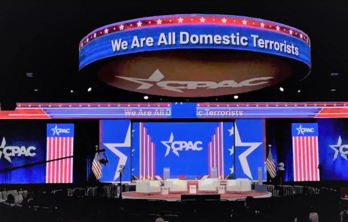 A stage at a CPAC event features a large circular sign above the stage that reads 'We Are All Domestic Terrorists'. The…