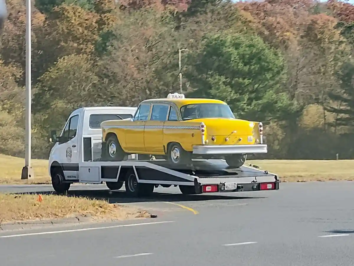 A white tow truck is hauling a vintage yellow taxi cab with a 'TAXI' sign on its roof along a road with autumn-colored trees…