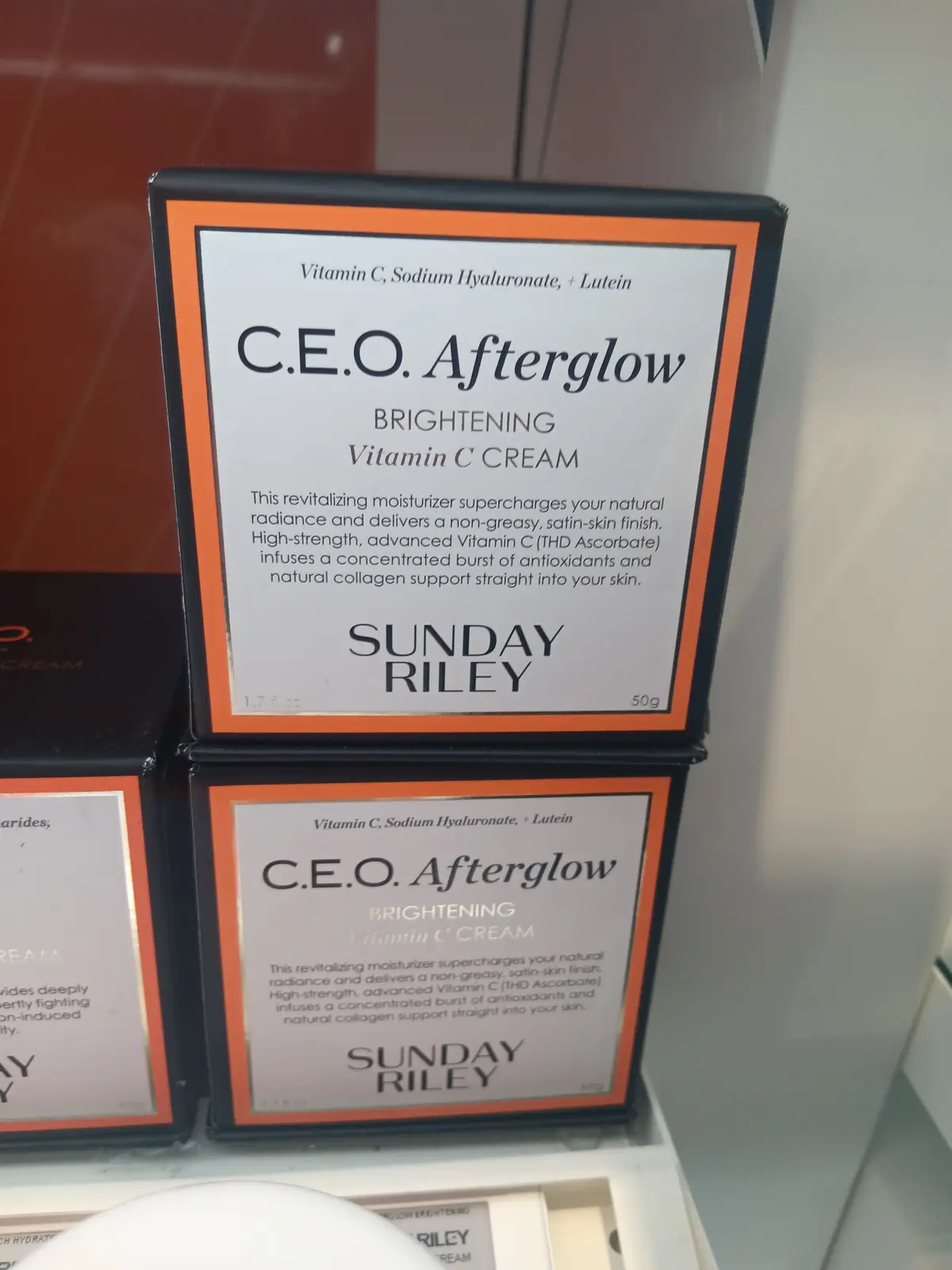 Two identical boxes of Sunday Riley's 'C.E.O. Afterglow Brightening Vitamin C Cream' are stacked on a shelf, with the…