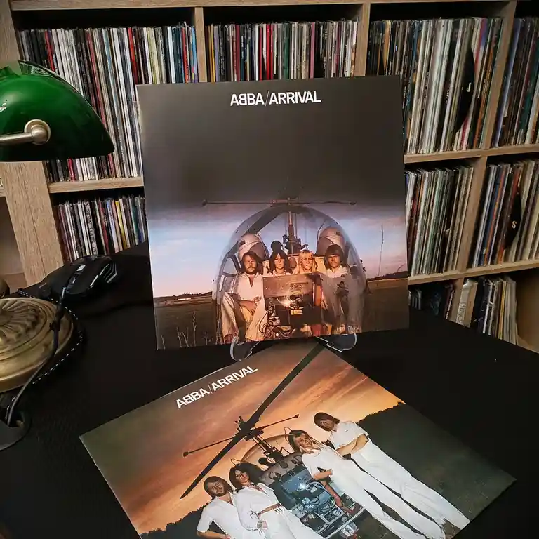 Two vinyl record album covers for ABBA's 'Arrival' are displayed on a desk, one upright and one flat, both featuring the…