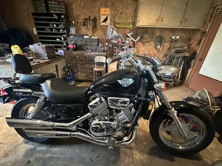 A black cruiser motorcycle with chrome accents is parked in a cluttered garage filled with tools, cabinets, and various…