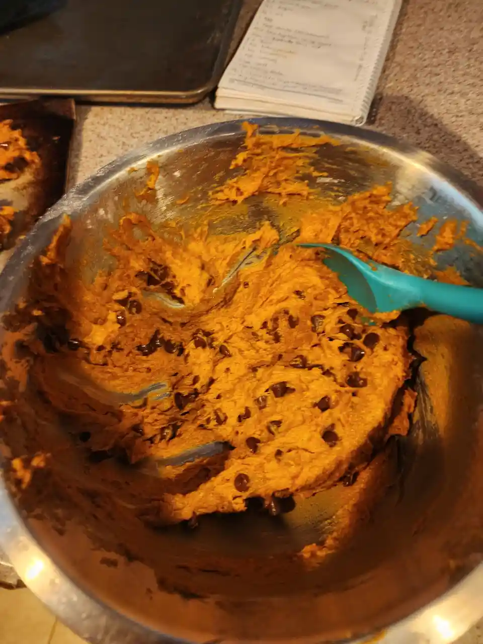 A close-up photo shows a metal mixing bowl containing thick, orange pumpkin chocolate chip cookie dough with a blue spatula…
