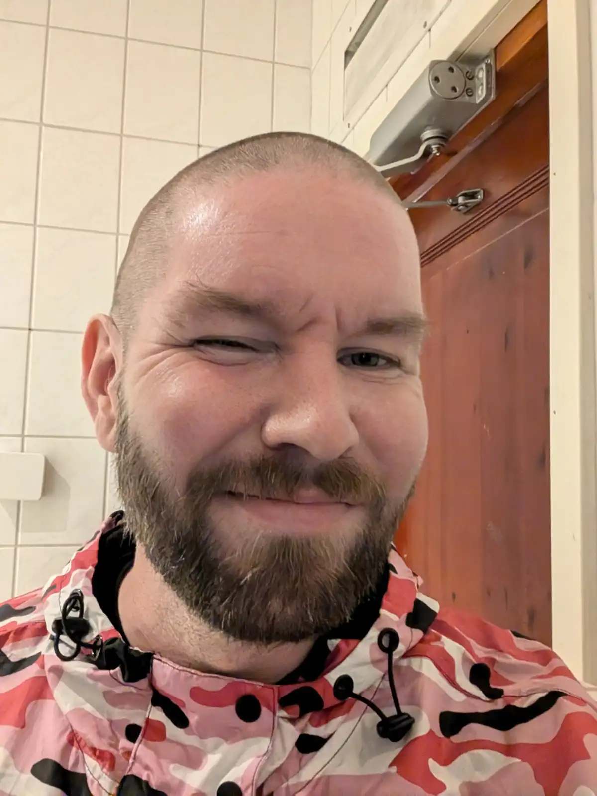 A man with a shaved head and beard, wearing a pink, white, and black camouflage-patterned jacket, makes a winking, smirking…