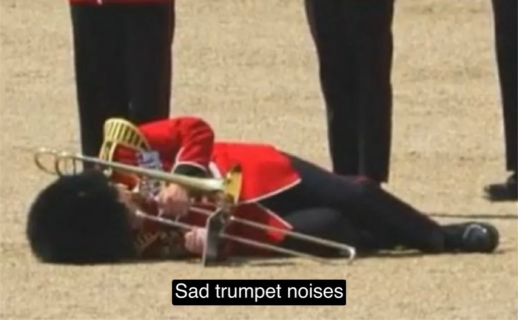 A person in a red uniform and black bearskin hat lies on the ground with a trombone, appearing to have fallen during a…