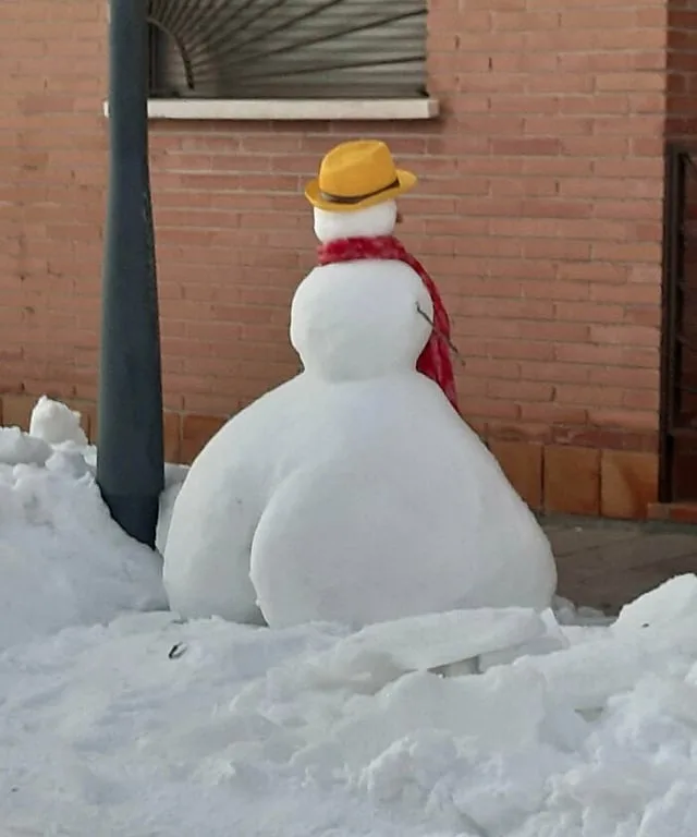 A snowman viewed from behind, wearing a yellow hat and red scarf, is sitting on a pile of snow with its back to the camera…
