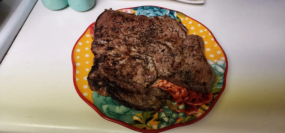 A large, seared steak rests on a colorful plate with a yellow polka-dot border and floral pattern, sitting on a white…