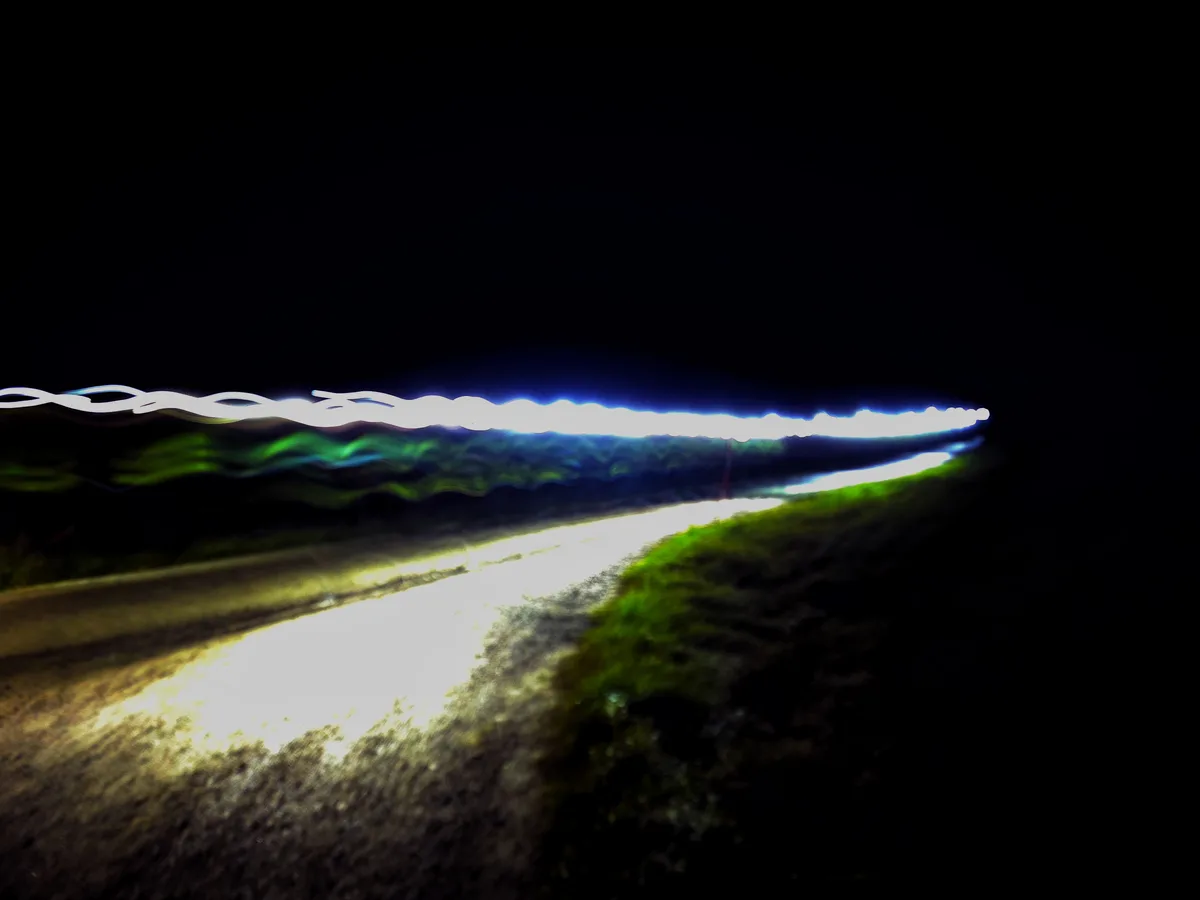 A long-exposure night photo shows a winding road illuminated by car headlights, with a bright, wavy white light streak above…