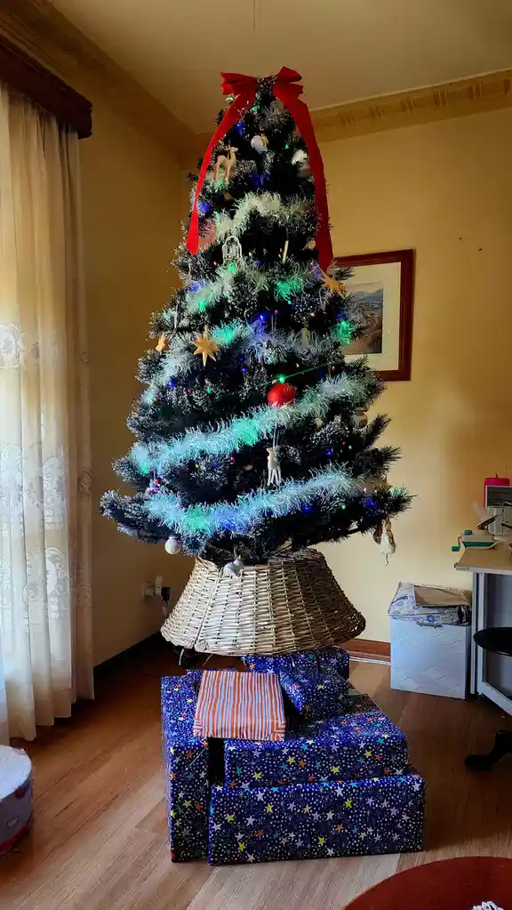 A decorated artificial Christmas tree with lights, ornaments, and a red bow is standing upright inside a large wicker…