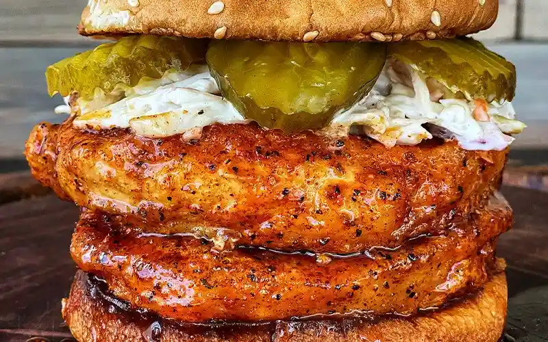 a close-up photo of a large chicken sandwich with two thick, glazed chicken patties stacked between buns, topped with…