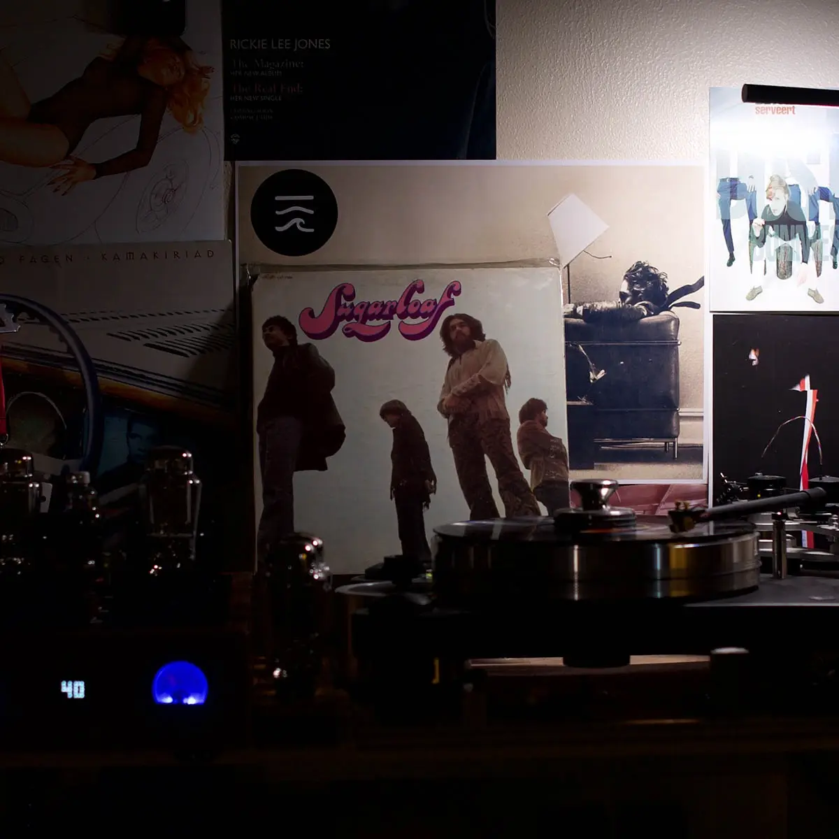 A dimly lit room features a vintage turntable with a vinyl record spinning, surrounded by various album covers on the wall…
