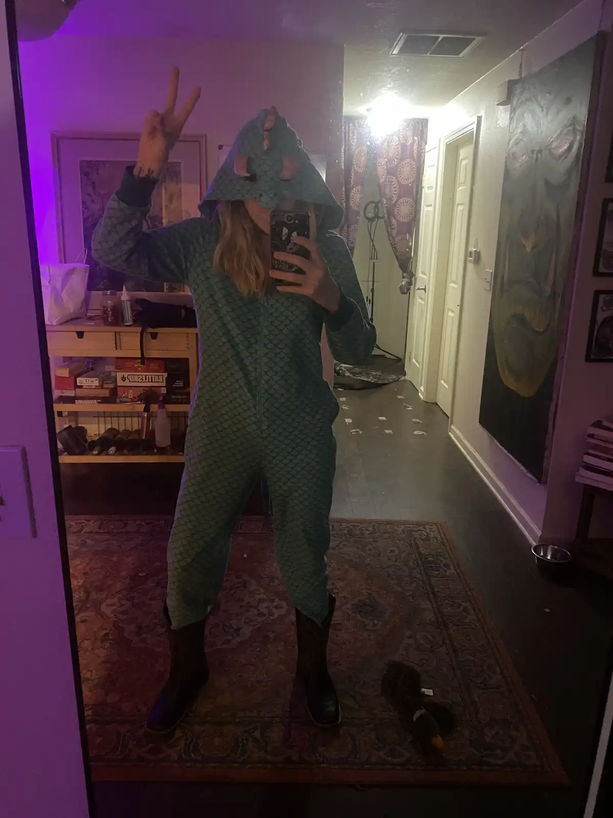 A person wearing a full-body teal onesie with a fish-scale pattern and a hood featuring fish-like fins and eyes is taking a…