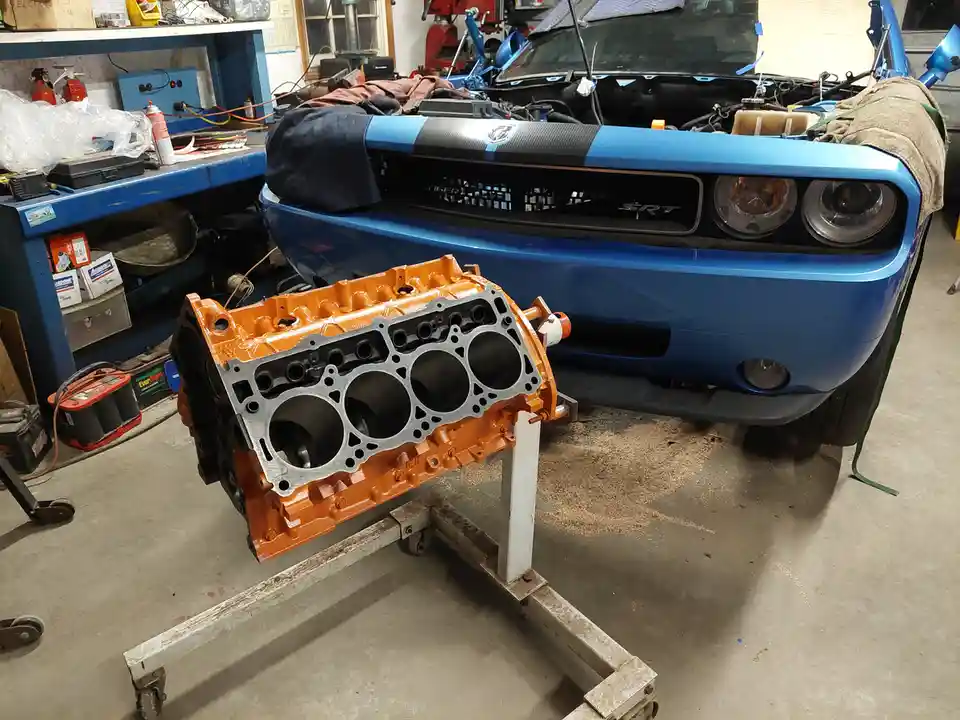 A blue Dodge Challenger SRT with its hood open sits in a cluttered garage, with a freshly painted orange engine block…