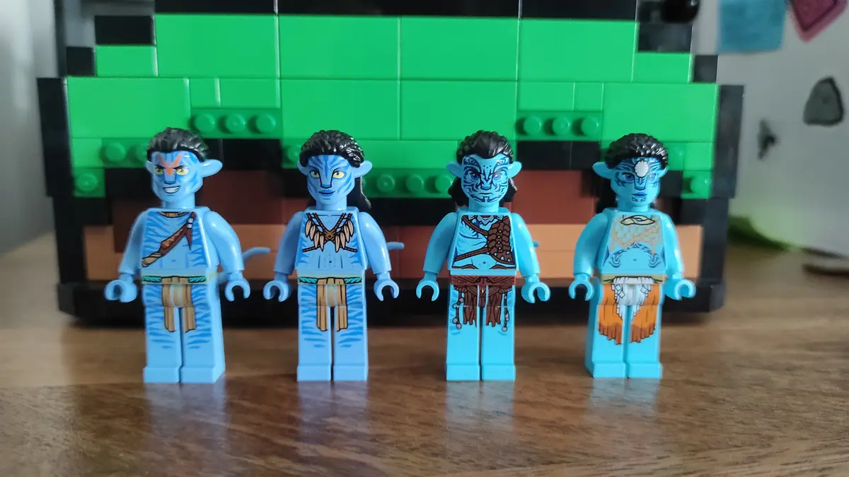 Four blue-skinned Avatar Na'vi LEGO minifigures stand in a row on a wooden surface, each with distinct facial markings and…