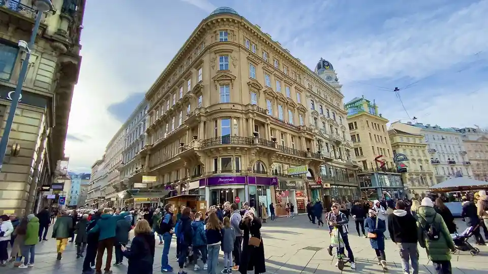 A wide-angle photo of a busy European city square with ornate, multi-story buildings and many pedestrians. A group of people…