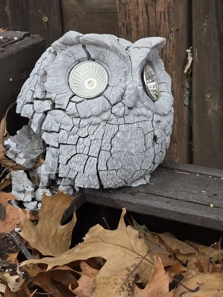 A weathered, gray owl-shaped garden light with cracked, peeling paint sits on a wooden surface surrounded by fallen brown…