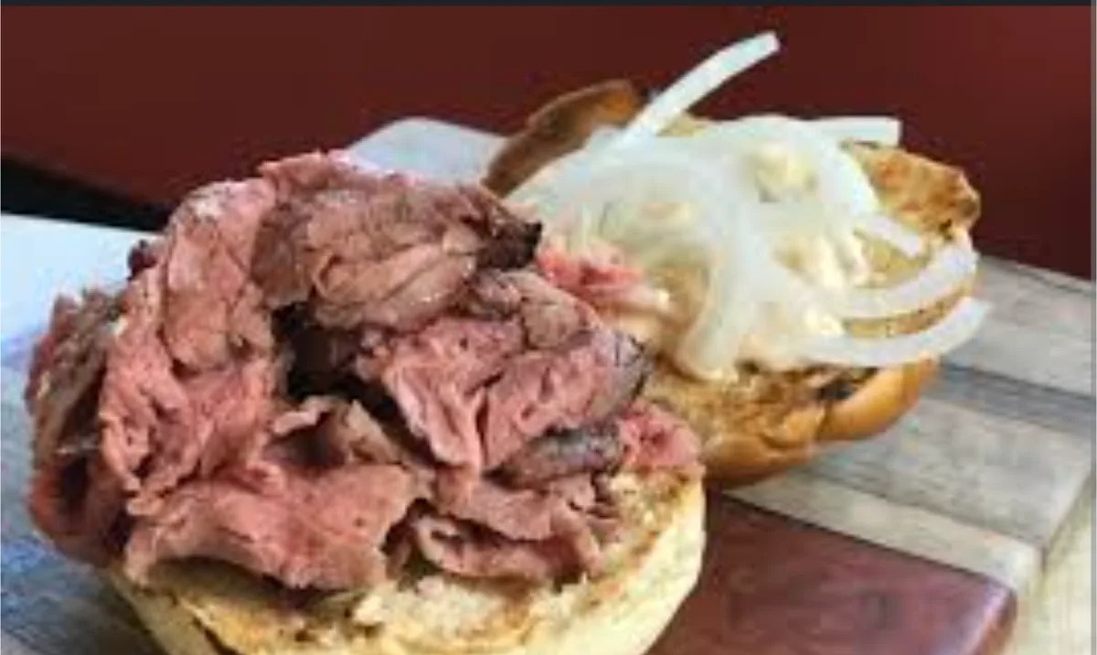 a close-up photo of a sandwich with a bottom bun piled high with shredded roast beef, and a top bun with sliced onions and a…