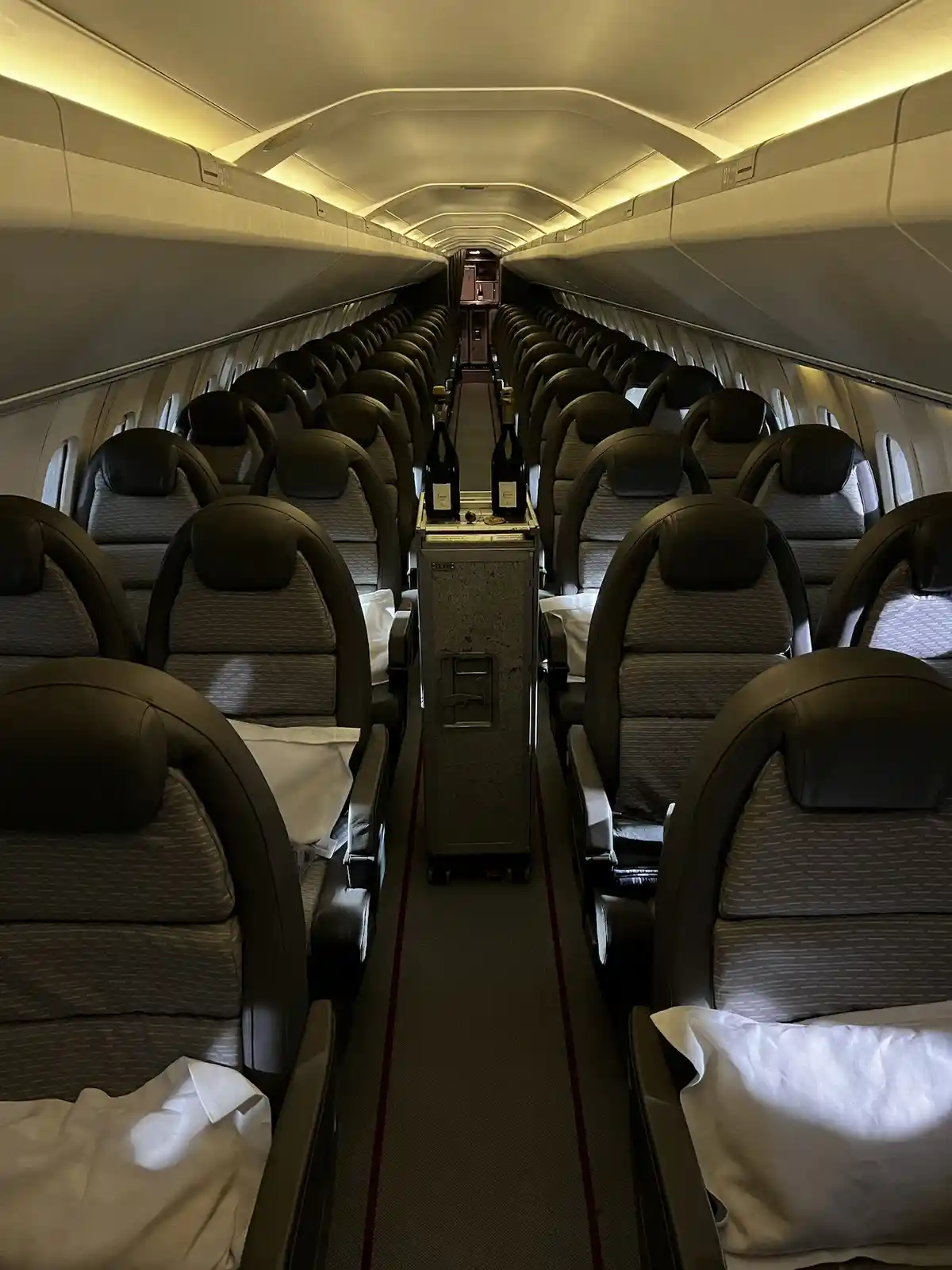 An empty airplane cabin with rows of unoccupied seats, white pillows on each, and a service cart with two champagne bottles…